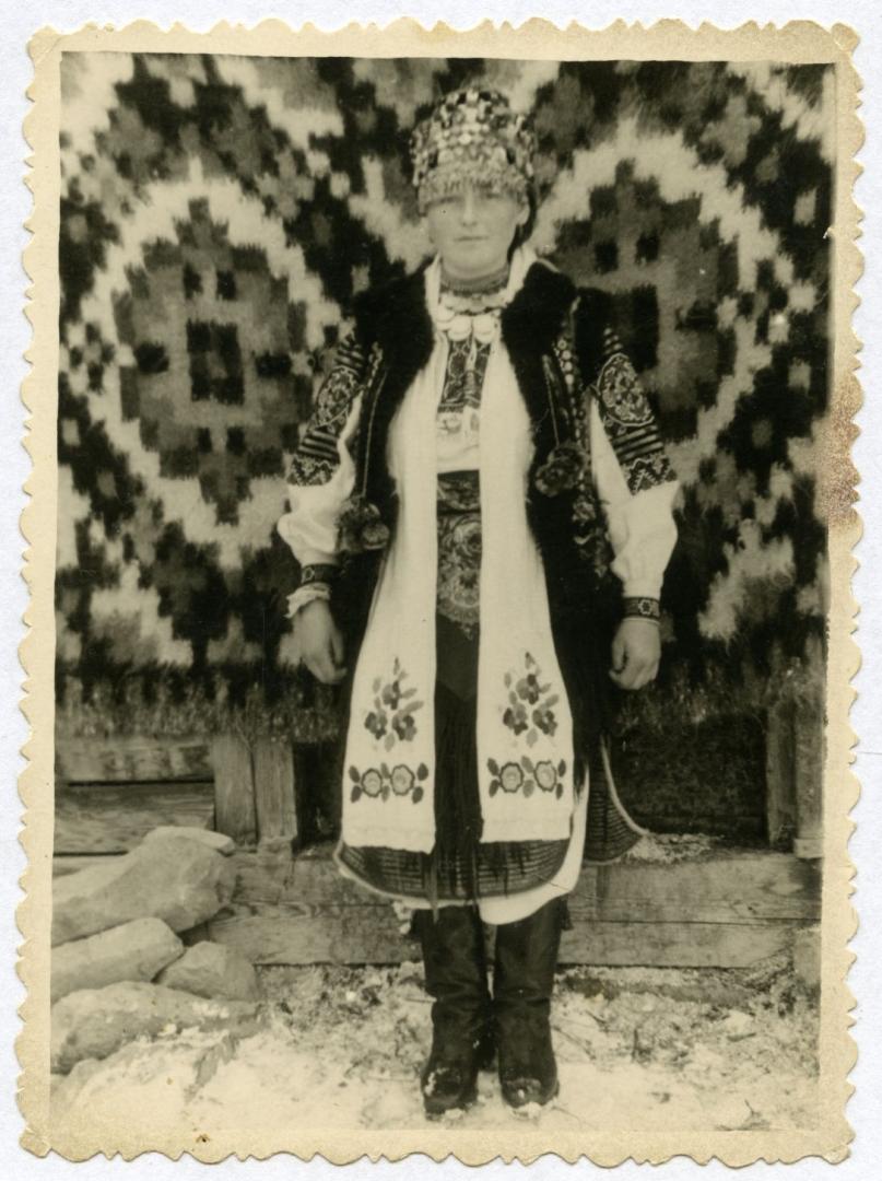 Photo. A girl wearing a folk wedding dress in front of a Hutsul lizhnyk (a thick, woolen bed cover or rug)