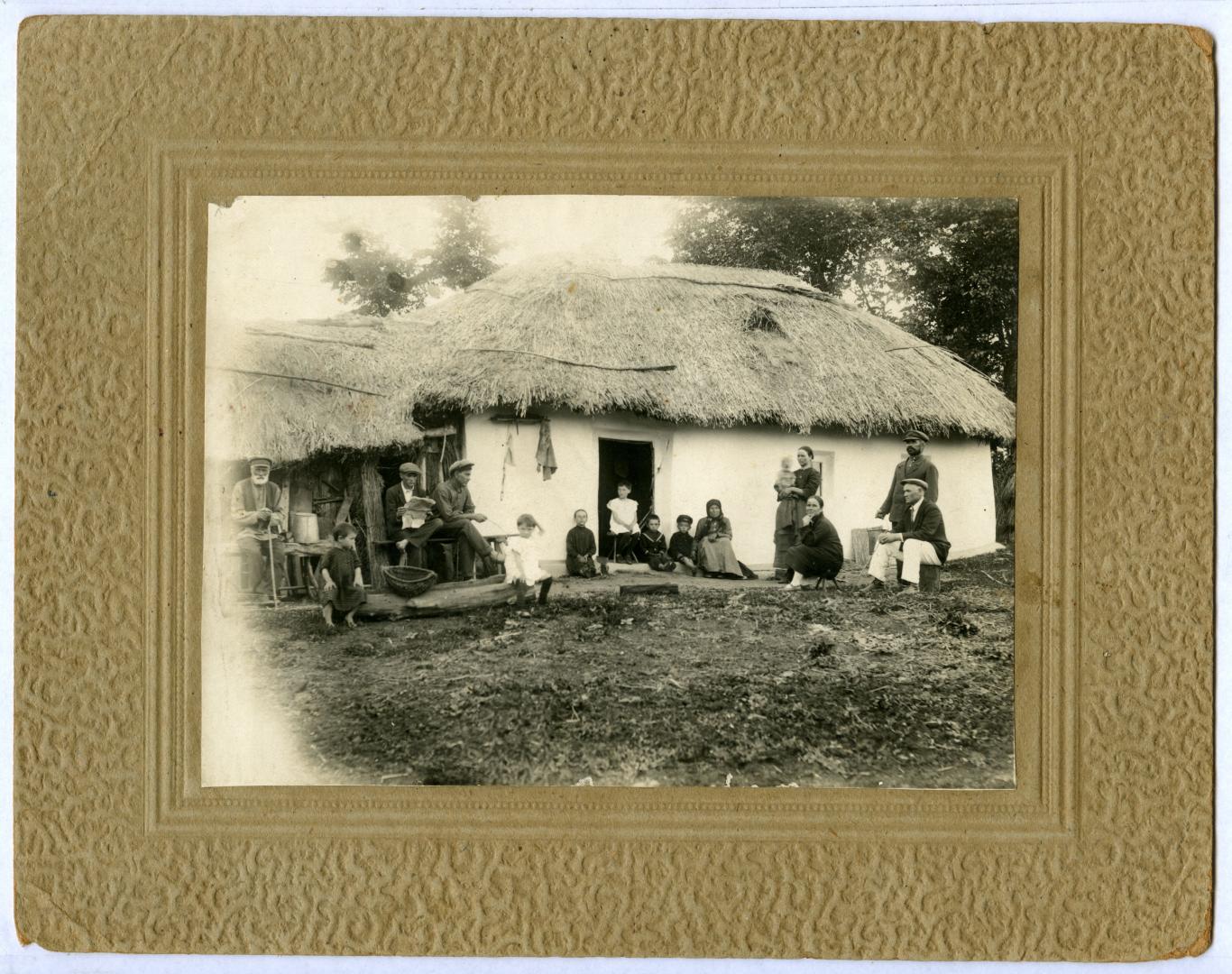 Photo. A family in front of a house, posed near a thatched roof and shed