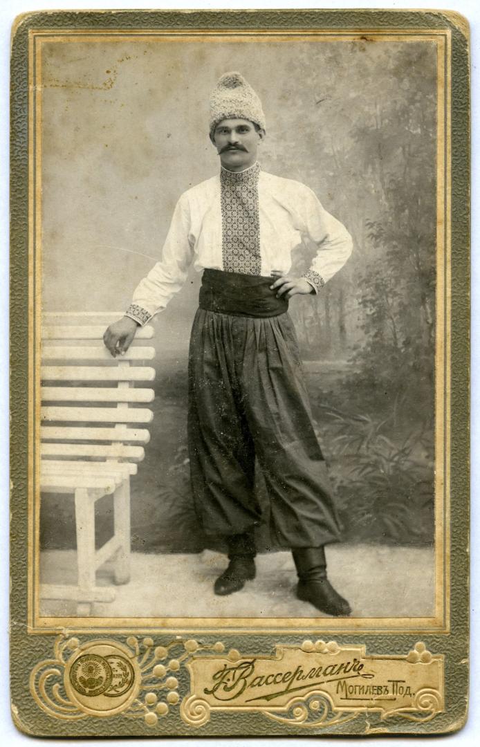 Photo. A young man wearing an embroidered shirt, sharovary (wide-legged pants), and a fur cap